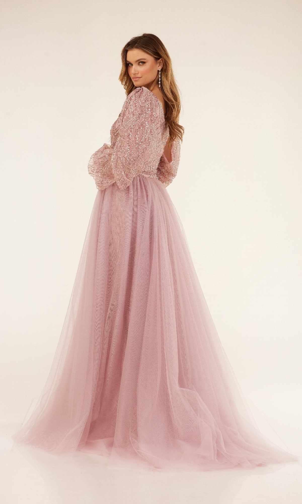 A woman models the Cecilia Couture C273 long formal dress, featuring a light pink hue, sheer sleeves, sequin details, and a flowing removable tulle overskirt against a plain light background.